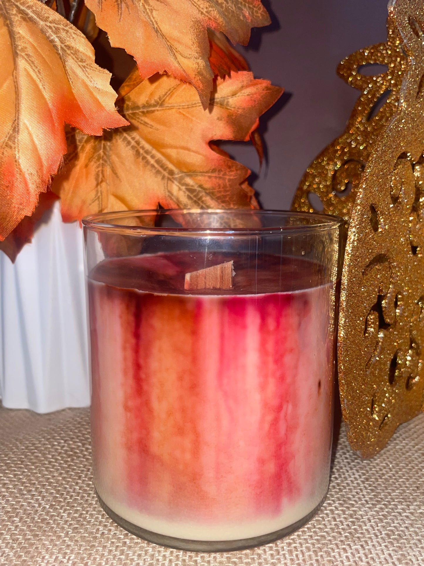 Candle in a glass container with beautiful vibrant fall coloured wax, a decorative leaf and gold ornament in the background