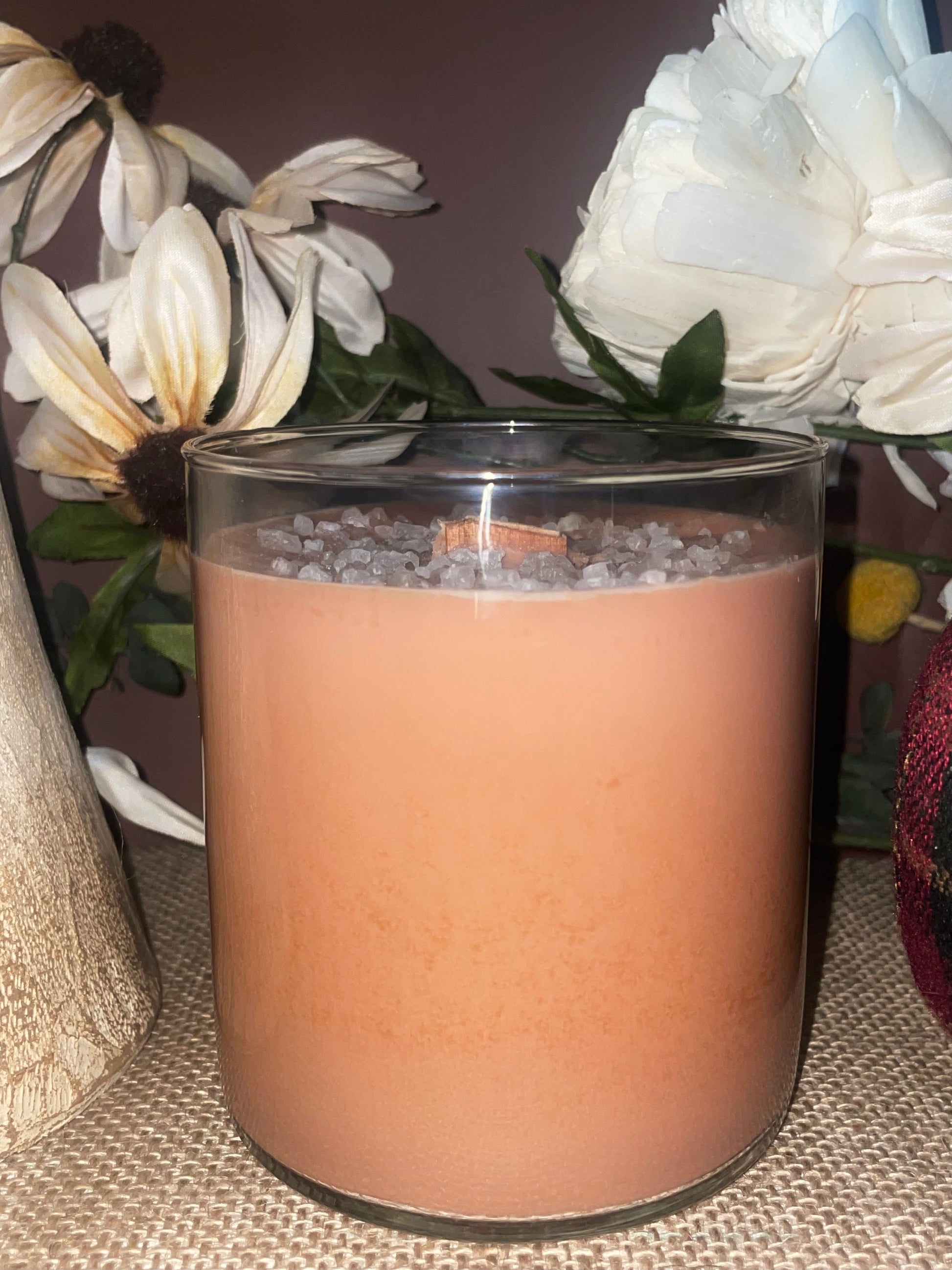 Candle in a glass jar on a textured wax surface with flowers in the background 