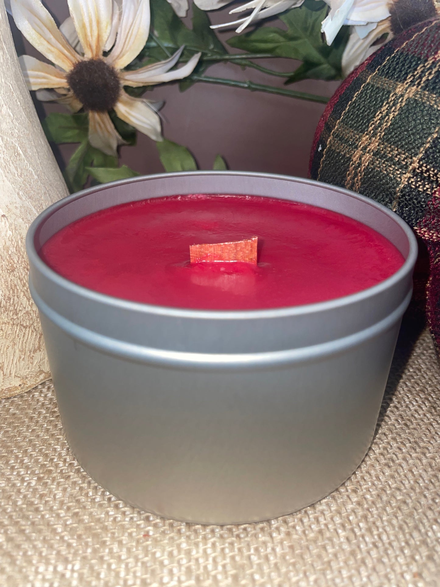 Red candle in a gray tin container on a textured surface with flowers in the background
