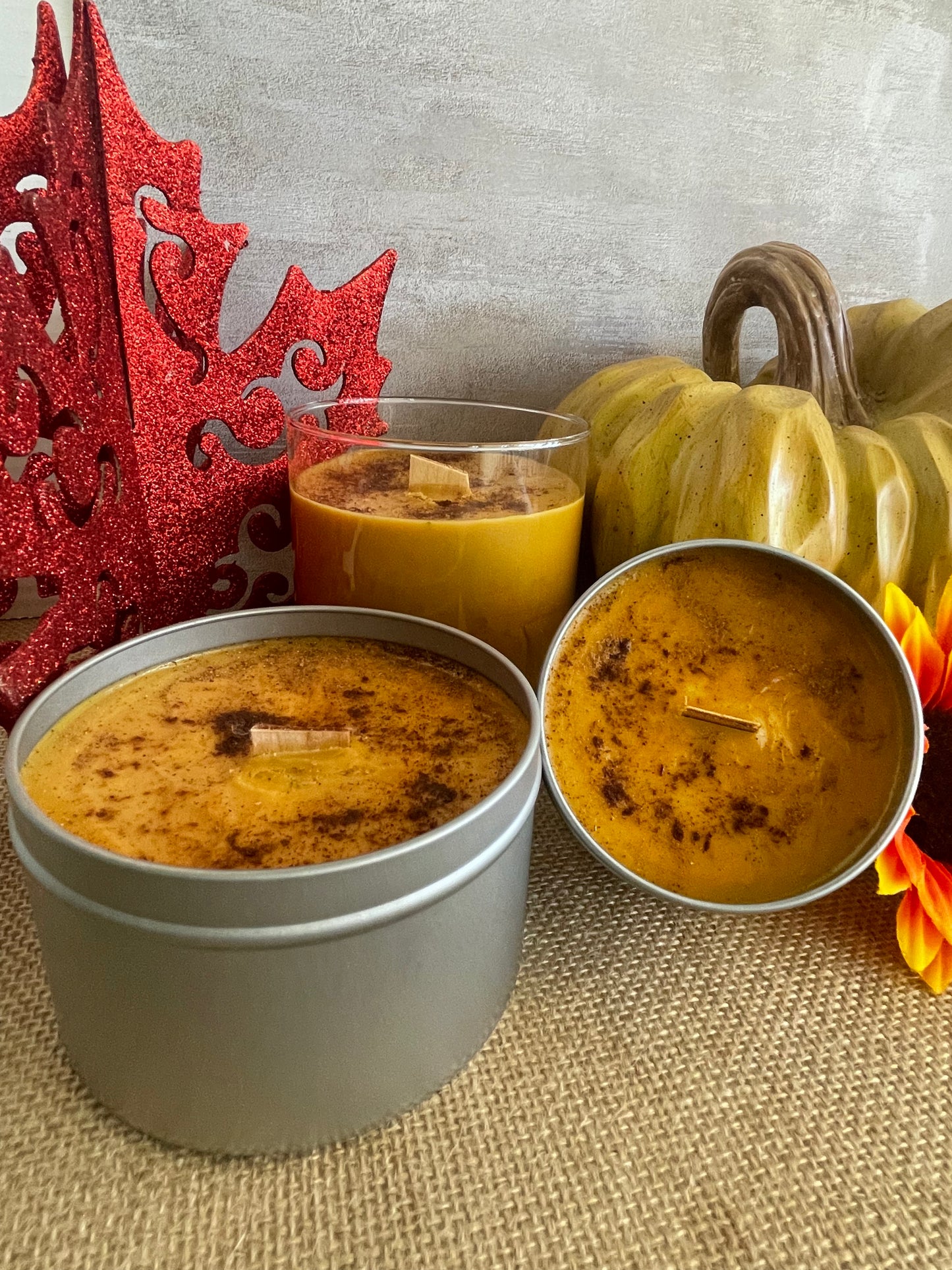 Three candles in containers with a decorative pumpkin and red fall decoration on a textured surface.