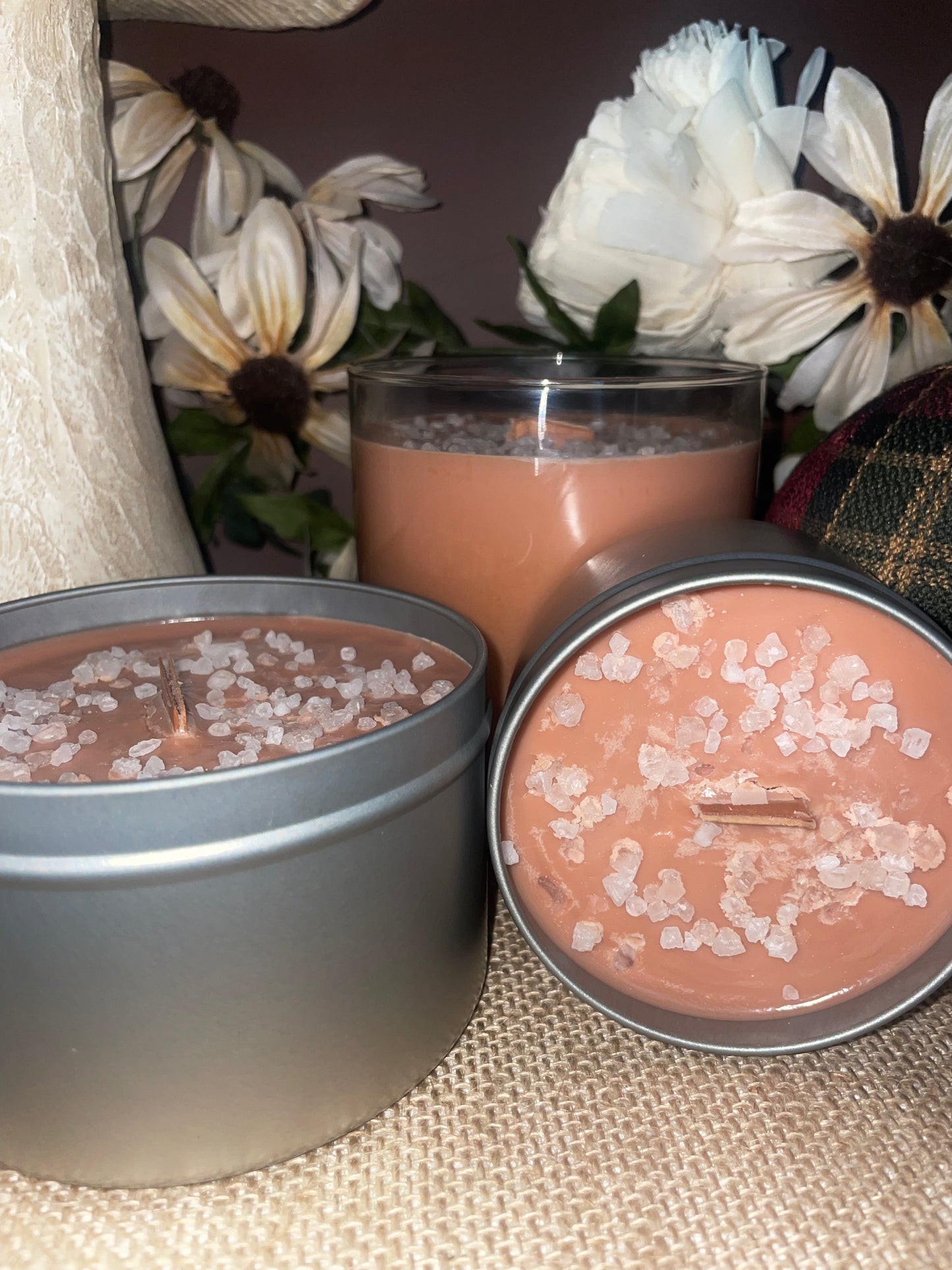Three candles, two in tins and one in a glass jar, with decorative flowers in the background.