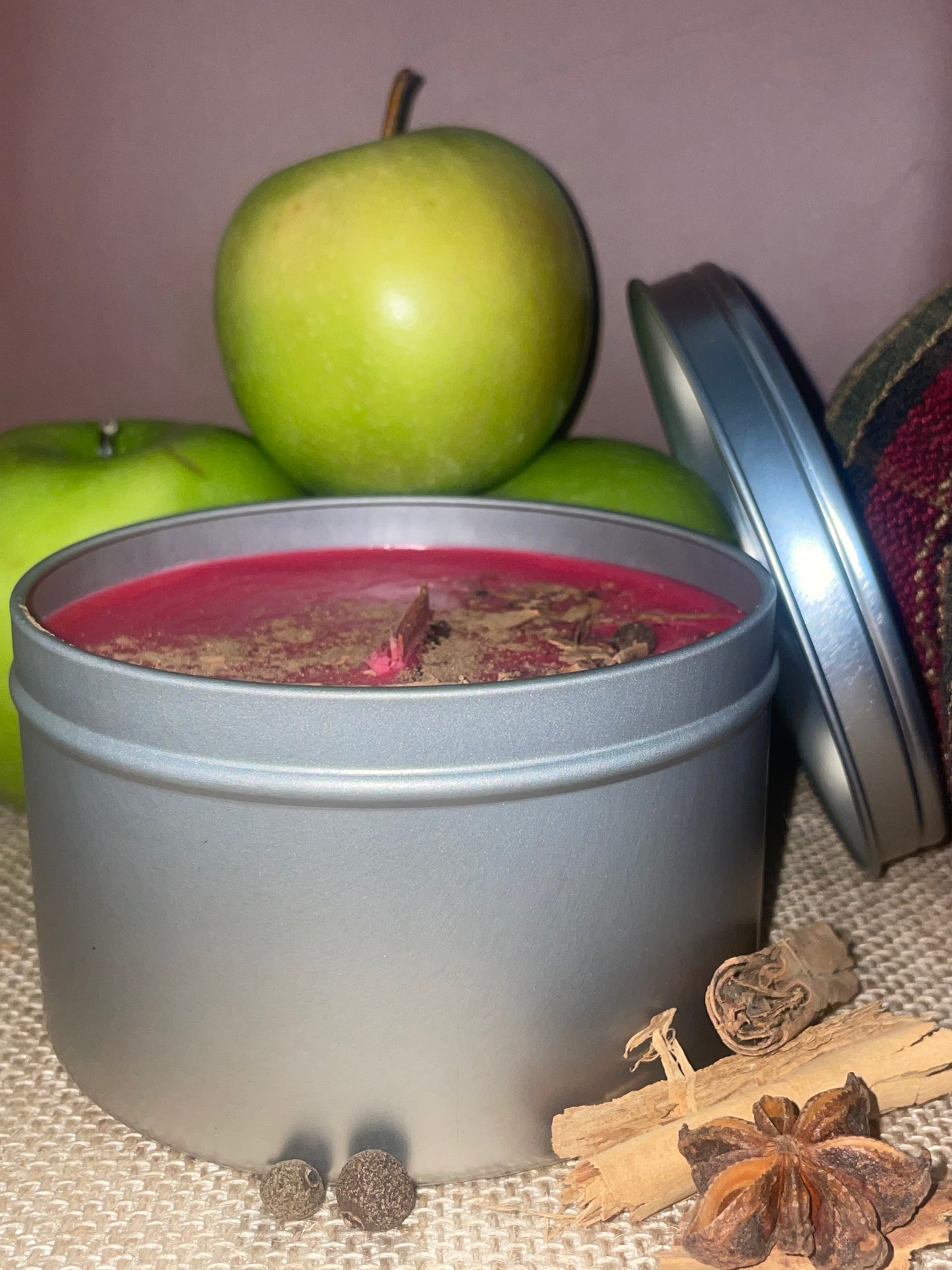 Silver tin with red candle inside, surrounded by green apples, cinnamon sticks, and star anise on a textured surface.